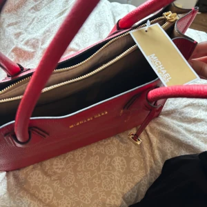 Röd handväska från Michael Kors - Snygg röd handväska från Michael Kors med guldfärgade detaljer och lås. Väskan har två handtag, dragkedja upptill och flera fack inuti. Perfekt accessoar för att lyfta din outfit.