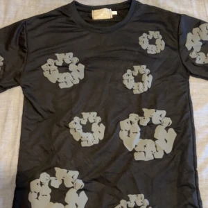 Denim tears T-shirt - Cool svart t-shirt från denim tears med unikt grått blommigt mönster över hela tröjan. Rund halsringning och korta ärmar. Perfekt för dig som vill sticka ut med en stilren men ändå lekfull look.