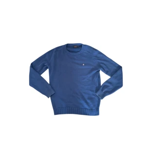 Blå stickad tröja från Morris - Snygg blå stickad tröja från Morris med rund halsringning och diskret broderad logga på bröstet. Tröjan har långa ärmar och ribbade muddar vid ärmslut och nederkant. Perfekt för lager-på-lager-stil. Storlek M. Passar mig på 185cm bra.