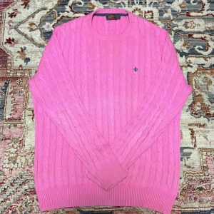 Rosa kabelstickad tröja från Morris - Säljer en rosa kabelstickad tröja från Morris i 100% pima bomull. Tröjan har rund halsringning, lång ärm och en liten broderad logotyp på bröstet. Perfekt för dig som gillar klassisk stil med färg.