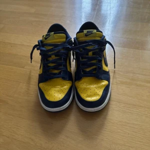 Nike Dunk Low blå och gula sneakers - Säljer ett par Nike Dunk Low sneakers i blått och gult med vit sula. Skorna har klassisk snörning och låg profil, perfekt för dig som gillar streetwear och vill sticka ut med färg. Ikonisk Nike-logga på sidan och plös.