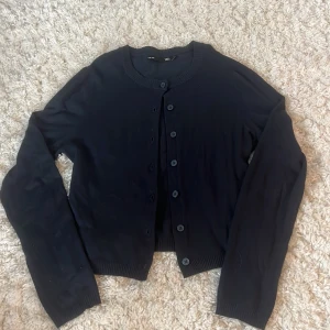 Svart kofta från 157 - Säljer en klassisk svart kofta från 157 i storlek S. Koftan har rund halsringning, långa ärmar och knäppning framtill med svarta knappar. Perfekt att slänga över en topp eller klänning. Mjuk och skön med ribbade muddar.