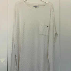 Vit långärmad tröja från Elvine - Säljer en vit långärmad tröja från Elvine i strukturerad bomull med rutigt mönster. Tröjan har en bröstficka med knappdetalj och rund halsringning. Perfekt för en stilren och avslappnad look.