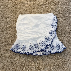 Vit omlottkjol med blå broderi och volang - Superfin vit kortkjol med omlott-design och volangkant. Kjolen är från H&M i storlek 36