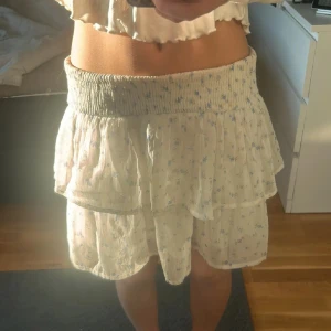 Vit blommig volangkjol med smock - Supersöt vit kortkjol med små blå blommor och bred smock i midjan. Kjolen har volanger och är gjord i ett lätt, luftigt material som passar perfekt till sommaren.