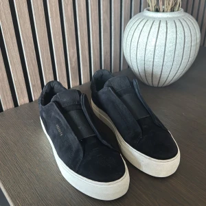 Svarta sneakers från Axel Arigato - Stilrena svarta sneakers från Axel Arigato med vit platt sula och diskret logga på sidan. Skorna är tillverkade i mocka och har en minimalistisk design utan synlig snörning, vilket ger en clean look. Köpare står för frakt 📦 