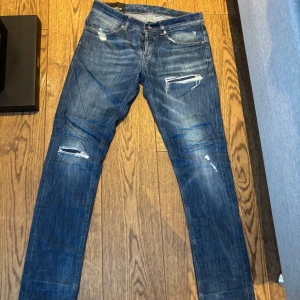 Blå slitna jeans från Dondup - Säljer ett par blå jeans från Dondup med slitna detaljer och raka ben. Jeansen har klassisk femficksdesign, dragkedja och knapp i gylfen samt snygga slitningar framtill för en trendig look.