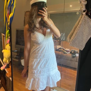 Vit spetsklänning med volang - Säljer en söt vit klänning med smala axelband och v-ringning. Klänningen har fina spetsdetaljer och volang nertill, samt en lätt utsvängd passform. Perfekt för studenten och sommarens alla tillfällen!