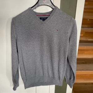 Grå v-ringad tröja från Tommy Hilfiger - Klassisk grå v-ringad tröja från Tommy Hilfiger med liten logga på bröstet. Tröjan har långa ärmar och ribbade muddar vid ärmslut och nederkant. Perfekt för lager-på-lager och stilrena outfits.