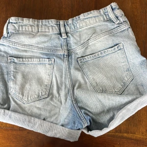 Ljusblå jeansshorts med upprullad kant - Säljer ett par ljusblå jeansshorts med upprullad kant och slitna detaljer framtill. Shortsen har klassiska fem fickor och bälteshällor. Perfekta för varma dagar och en avslappnad stil.