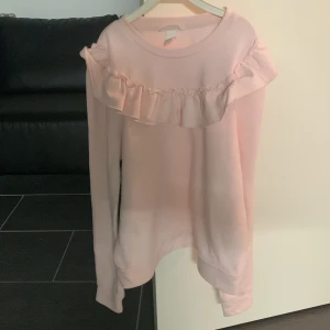 Ljusrosa sweatshirt med volang - Säljer en ljusrosa sweatshirt med rund hals och volangdetalj över bröstet och axlarna. Tröjan har långa ärmar och är i mjukt material, perfekt för en söt och avslappnad stil.