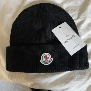 Svart stickad mössa från Moncler - Svart ribbstickad mössa från Moncler med uppvikt kant och Moncler-logga framtill. Klassisk och stilren design som passar perfekt till kyliga dagar. Säljs pågrund gillar den inte mer. Skriv för minsta fundering.