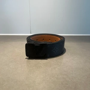 Svart LV bälte - Säljer ett svart bälte från Louis Vuitton med klassiskt LV-monogram och svart spänne. Bältet har en brun insida och är gjort i skinn. Perfekt accessoar för att lyfta din outfit.