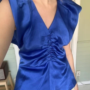 Blå satinblus med v-ringning och rynkdetalj - Säljer en snygg blå blus i satin med v-ringning och rynkdetalj framtill. Blusen har korta ärmar med lätt puff och en figurnära passform. Perfekt för dig som vill sticka ut med en färgstark topp! Säljer då den är lite för stor på mig 💙🩵