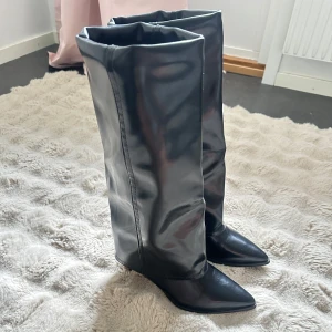 Svarta stövlar med klack och spetsig tå - Säljer ett par höga svarta stövlar i glansigt syntetmaterial med spetsig tå och smal klack. Stövlarna har en slouchy passform och når upp till knäna. Perfekta för att lyfta din outfit. Jättefina, trendiga boots/ stövlar med vik, som är knähöga eller precis under knäna rättare sagt (går att se på sista bilden) Använda en gång, inomhus❤️❤️ klacken är ej hög typ 5cm❤️ storlek 37