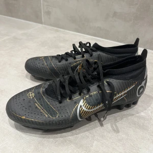 Svarta Nike fotbollsskor med guldiga detaljer - Säljer ett par svarta Nike fotbollsskor med snygga guldiga och vita detaljer. Skorna har snörning och en platt sula med dobbar för bra grepp på planen. Perfekta för dig som vill sticka ut på fotbollsplanen. 