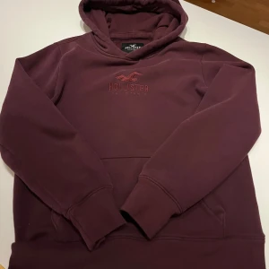 Vinröd hoodie från Hollister - Mysig vinröd hoodie från Hollister med broderad logga på bröstet. Tröjan har huva och en stor magficka framtill. Perfekt för en avslappnad och stilren look.