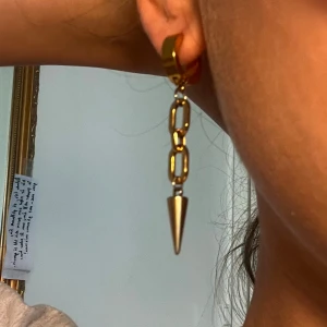 Guldiga kedjeörhängen med konformad detalj - Säljer ett par statement örhängen i guld med kedjelänkar och en konformad hängande detalj. Snygga och trendiga örhängen som verkligen drar blickarna till sig.