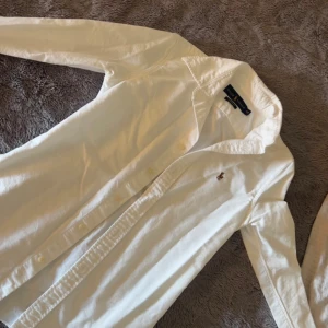Vit skjorta från Ralph Lauren - Klassisk vit skjorta från Ralph Lauren med broderad logga på bröstet. Skjortan har lång ärm, krage och knäppning framtill. Perfekt för en stilren och tidlös look.