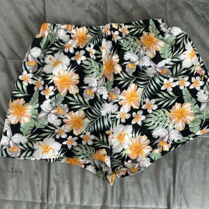 Blommiga shorts med tropiskt mönster - Färgglada shorts med tropiskt blommönster i gult, vitt och grönt på svart botten. Perfekta för sommaren med avslappnad passform och elastisk midja. Lätta och bekväma, passar till strand eller varma dagar.