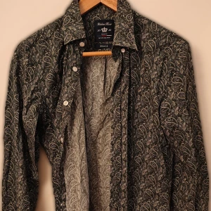 Mönstrad skjorta från Robbie Moor - Säljer en grön långärmad skjorta från Robbie Moor i slim fit-modell. Skjortan har ett detaljerat paisleymönster i grönt och vitt samt knappar framtill. Perfekt för dig som vill sticka ut med en unik stil.