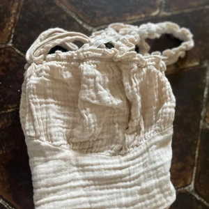 Beige croppad linnetopp med volangdetaljer - Säljer en söt beige croppad linnetopp med volangdetaljer och smala axelband. Toppen har en rynkad struktur och knyt framtill. Perfekt för varma dagar och enkel att styla till olika outfits.