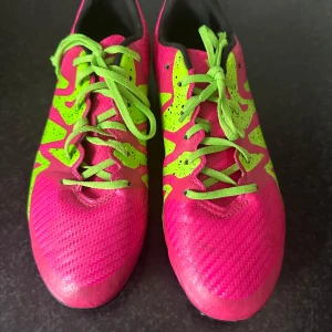 Adidas fotbollsskor i rosa och neongrönt - Säljer ett par Adidas fotbollsskor i en riktigt snygg rosa färg med neongröna detaljer och snörning. Skorna har svart sula med dobbar och är perfekta för dig som vill sticka ut på planen.