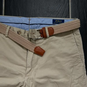 Beige chinos från Polo Ralph Lauren med bälte - Snygga beige chinos från Polo Ralph Lauren i slim fit-modell. Byxorna har en klassisk design med fickor fram och bak, samt ett medföljande flätat bälte i beige och brunt. Perfekta för en stilren och avslappnad look. Bälte följer med därav priset! 