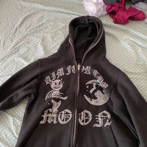 Aim for the moon kofta - Svart hoodie med dragkedja och huva. Framsidan har stora motiv i strass, bland annat en tecknad figur och text. Hoodien har en avslappnad passform och är perfekt för en streetwear-look.