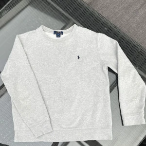 Grå sweatshirt från Polo Ralph Lauren - Säljer en klassisk grå sweatshirt från Polo Ralph Lauren med rund halsringning och den ikoniska lilla broderade loggan på bröstet. Tröjan har långa ärmar och är tillverkad i mjuk bomull. Perfekt för en avslappnad stil. 14-16 passar S.