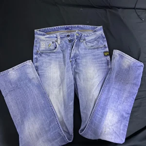 Blå jeansbyxor från G-Star RAW - Säljer ett par klassiska blå jeans från G-Star RAW med raka ben och snygga slitningar. Byxorna har dragkedja på bakfickan, detaljerade sömmar och G-Star-logga på bakfickan. Perfekta för dig som gillar streetwear och vill ha en cool look.