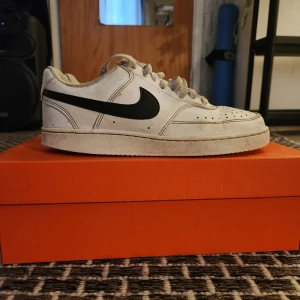 Nike courtage vision - Klassiska vita Nike Air Force 1 sneakers med svart swoosh. Skorna har snörning, rund tå och platt sula. Perfekta för dig som gillar en tidlös och sportig stil. Säljs i original orange Nike-låda.