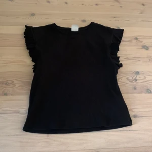 Svart ribbad topp med volangärmar från Zara - Säljer en svart ribbad topp från Zara i storlek 152. Toppen har korta ärmar med volangdetaljer som ger en söt touch. Perfekt att matcha med jeans eller kjol för en stilren look.