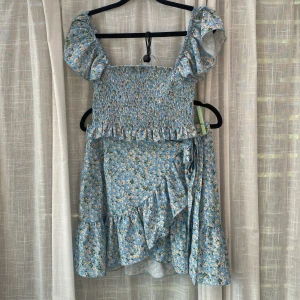 Blommig blå sett med volang - Söt blå sett med topp och kjol. Har små vita blommor, volangdetaljer vid axlar och nederkant samt smockad överdel. Klänningen har breda axelband och omlottkänsla på kjolen. Perfekt för sommarens varma dagar.