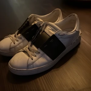 Valentino Open sneakers i vitt och svart - Säljer ett par vita Valentino Open sneakers med svart band över vristen och ikoniska nitar på hälen. Skorna har rund tå, platt sula och snörning. Perfekta för dig som gillar stilrena och lyxiga sneakers med en twist. Enligt skorna hänger med. Pris kan diskuteras! Nypris 7300kr.