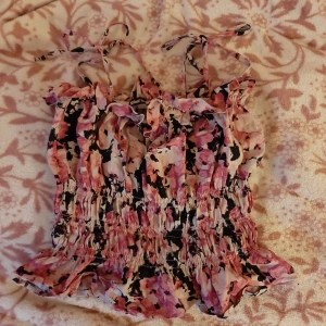 Blommig Tub top från Gina Tricot (Med volanger) - Supersöt blommig topp från Gina Tricot med volangdetaljer och smockad midja. Toppen har tunna axelband som knyts och ett färgglatt mönster i rosa, svart och vitt. Perfekt för varma dagar och ger en romantisk känsla.