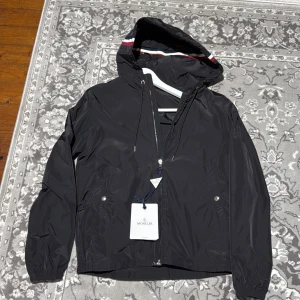 Svart vindjacka från Moncler med huva - Snygg svart vindjacka från Moncler med huva och dragkedja. Jackan har Moncler-logga på ärmen och en diskret röd och vit rand på huvan. Perfekt för blåsiga dagar och enkel att matcha med olika outfits.