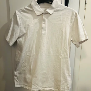 Vit pikétröja från H&M - Klassisk vit pikétröja från H&M med krage och knappar framtill. Tillverkad i mjuk bomull och har korta ärmar. Perfekt för en stilren och enkel look.