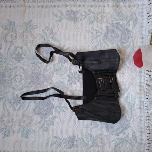 Svart axelväska med spännen - Säljer en svart axelväska i syntetmaterial med justerbar axelrem och dekorativa spännen framtill. Väskan har en rektangulär form och flera fack för enkel organisering. Perfekt för dig som vill ha en stilren och praktisk accessoar.