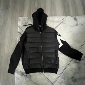 Svart pufferjacka med huva och vit detalj - Svart pufferjacka med huva och dragkedja framtill. Jackan har quiltade partier på framsidan och en stor vit pil-detalj på ena ärmen. Mjuka muddar vid ärmslut och nederkant. Pris kan diskuteras! 