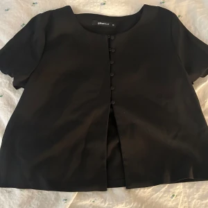 Svart kortärmad kavaj från Gina Tricot - Stilren svart topp från Gina Tricot med korta ärmar och rund halsringning. Kavajen har dekorativa knappar framtill och en rak, något croppad passform. Endast använd 1 gång