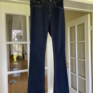 Mörkblå bootcut jeans från b.young - Säljer ett par mörkblå jeans från b.young med klassisk bootcut-modell. Jeansen har fem fickor, synliga sömmar och stängs med dragkedja och knapp. Perfekta för dig som gillar en tidlös och stilren look.
