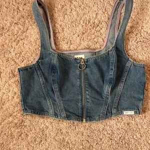 Jeans korsettopp med dragkedja från Lee x H&M - Säljer en snygg jeans korsettopp från Lee x H&M. Toppen är croppad med breda axelband och har en silverfärgad dragkedja framtill med ringdetalj. Perfekt till sommaren och enkel att styla till olika looks. Skulle gissa att den passar storlek M och mindre. Endast använd en gång.