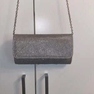 Glittrig silver balväska - Säljer en snygg kuvertväska i silver med glittrigt material och kedjerem. Perfekt för att lyfta din outfit! Väskan har magnetlås och ett fack inuti för småsaker. Använt en gång.