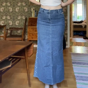 Lång jeanskjol med broderi - Säljer en lång jeanskjol från Eagame med ljusblått broderi längs sidorna. Kjolen har klassiska jeansfickor fram och bak, hög midja och stängs med dragkedja och knapp. Perfekt för dig som gillar denim med vackra detaljer.