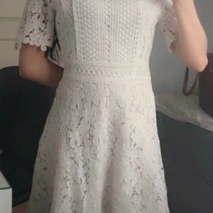 Vit spetsklänning med blommigt mönster - Superfin vit klänning i spets med blommigt mönster och korta ärmar. Klänningen har en markerad midja, rund halsringning och vackra detaljer framtill. Perfekt för sommarens tillfällen eller studentfirande. Säljes då den inte längre kommer till användning. Sitter jättefint på kroppen. Ej använd bara provats en gång.