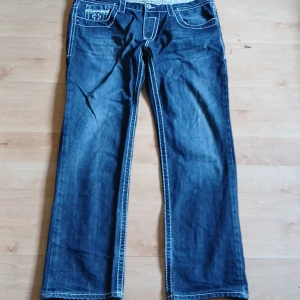 Mörkblå jeans med vit brodyr - Säljer ett par mörkblå jeans med tydliga vita kontrastsömmar och broderade detaljer på bakfickorna. Klassisk femficksmodell med raka ben och knappgylf. Snygga detaljer och unik look.