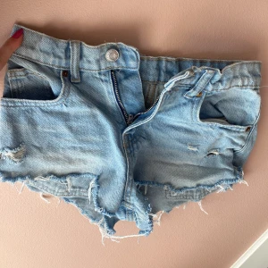 Ljusblå jeansshorts från Zara - Säljer ett par ljusblå jeansshorts från Zara med råa, fransiga kanter och slitna detaljer. Klassisk femficksmodell med låg midja och dragkedja. Perfekta för sommaren och riktigt snygga till en enkel topp.