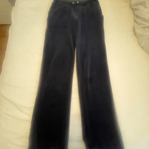Mörkblå velourbyxor från Juicy Couture - Mörkblåa velourbyxor från Juicy Couture i storlek XS. Byxorna har raka ben, resår i midjan med dragsko och två fickor med lock och knappar bak. Perfekta för en avslappnad och trendig look.Snöret finns men är inte isatt – går att fixa. Därav lägre pris