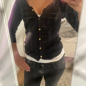 Svart kofta med guldknappar från H&M - Säljer en svart kofta från H&M i storlek XS. Koftan har dekorativa guldknappar framtill och ett diskret hålmönster över hela plagget. Perfekt att slänga över en topp för en snygg lager-på-lager-look.